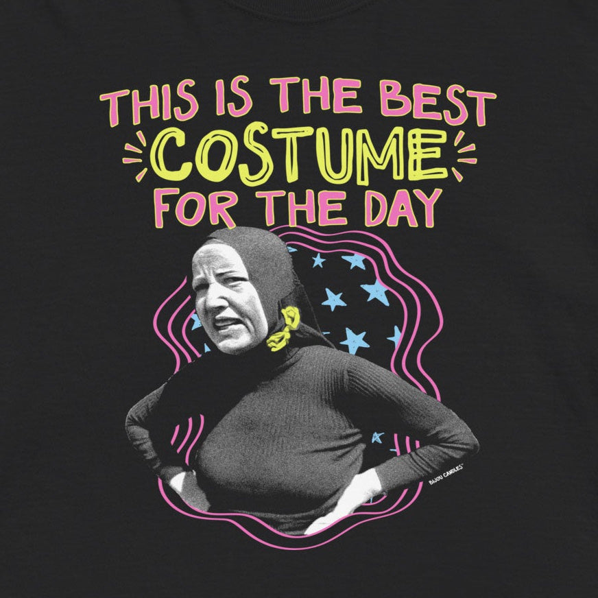 A black short-sleeve graphic t-shirt featuring a colorful print with the text 'THIS IS THE BEST COSTUME FOR THE DAY' and a stylized portrait of a person in a costume, displayed against a light cream background.