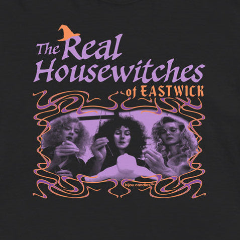A black short-sleeve graphic t-shirt featuring the text 'The Real Housewives of Eastwick' in purple and orange, with a central purple-toned illustration of three women, laid flat against a light cream background.