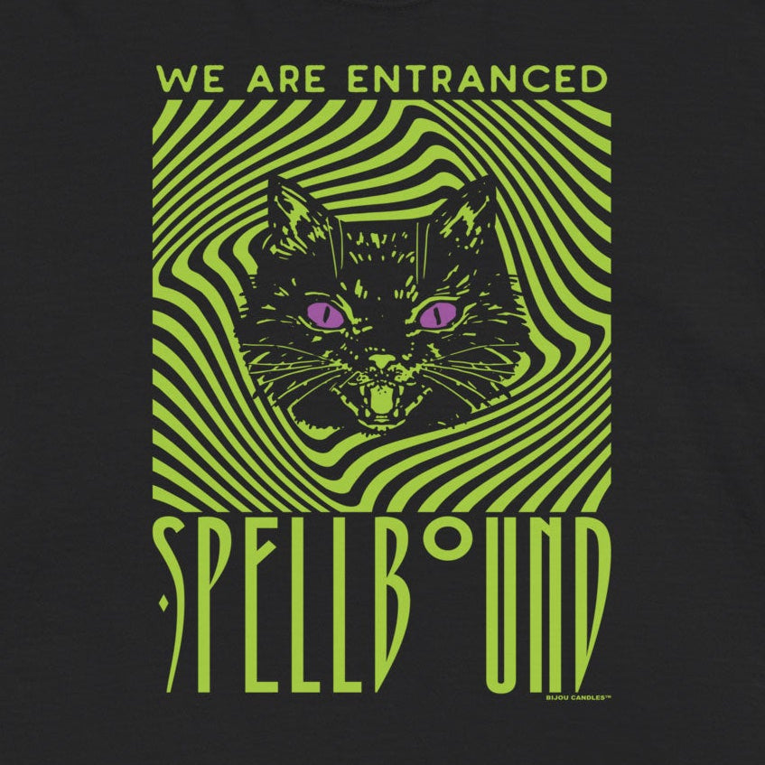 A black short-sleeve t-shirt with a green and black psychedelic graphic of a cat's face inside a swirling pattern, with the text 'WE ARE ENTRANCED' above and 'SPELLS UNBOUND' below, displayed on a light cream background.