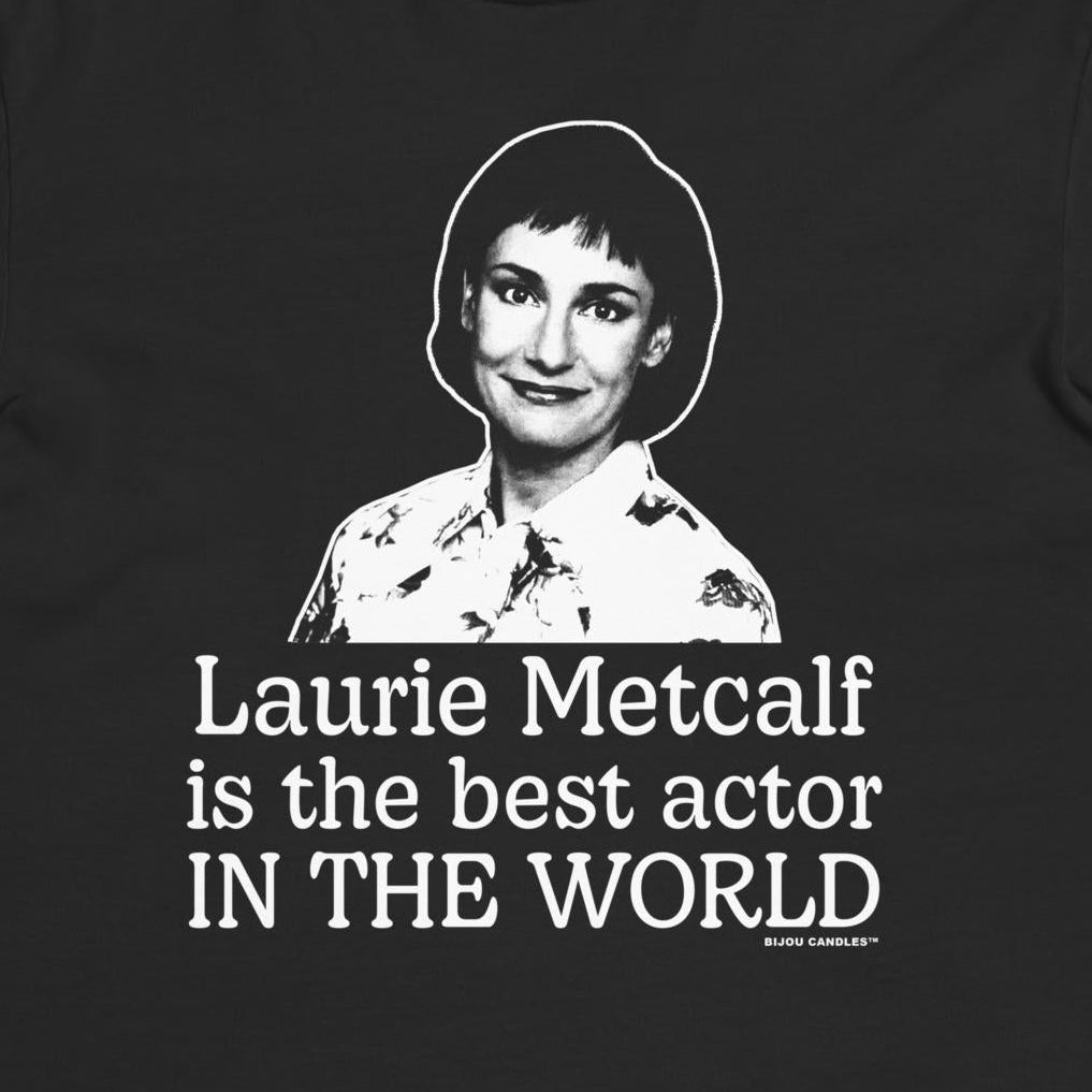 A black short-sleeve crewneck t-shirt featuring a white graphic print of a portrait of Laurie Metcalf and the text 'Laurie Metcalf is the best actor IN THE WORLD' on a light cream background.