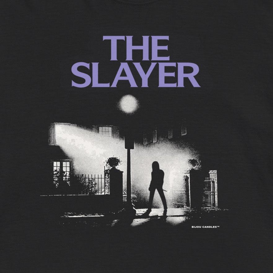 A black short-sleeve graphic t-shirt featuring the purple text 'THE SLAYER' above a black and white film still print, displayed flat against a light cream background.