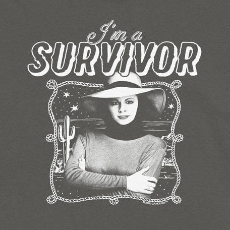 A dark gray short-sleeve t-shirt with a black and white graphic print on the front. The graphic features the text 'I'm a SURVIVOR' above a vintage-style portrait of a woman wearing a cowboy hat.