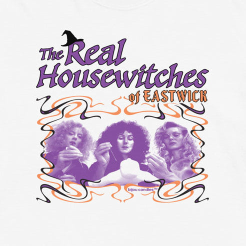 A white short-sleeve t-shirt with a graphic print of 'The Real Housewives of Eastwick' featuring purple and orange tones, displayed flat against a light cream background.